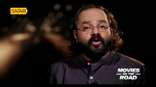 A man with a beard and glasses speaks directly to the camera, his mouth slightly open. The Safari TV logo appears in the upper left corner, and a "Movies on the Road" graphic is visible on his shoulder.