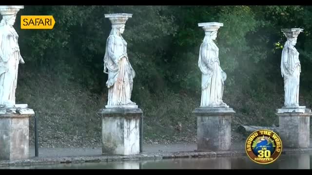 A line of white marble statues stands before a dense green backdrop. A small, brown bird hops across the path behind the third statue from the left.