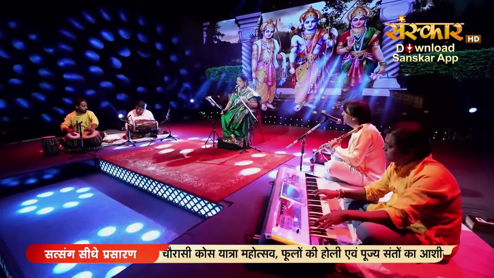 A woman in a green sari sings into a microphone while a man plays a flute beside her. To their left, two musicians tap out rhythms on tabla drums.