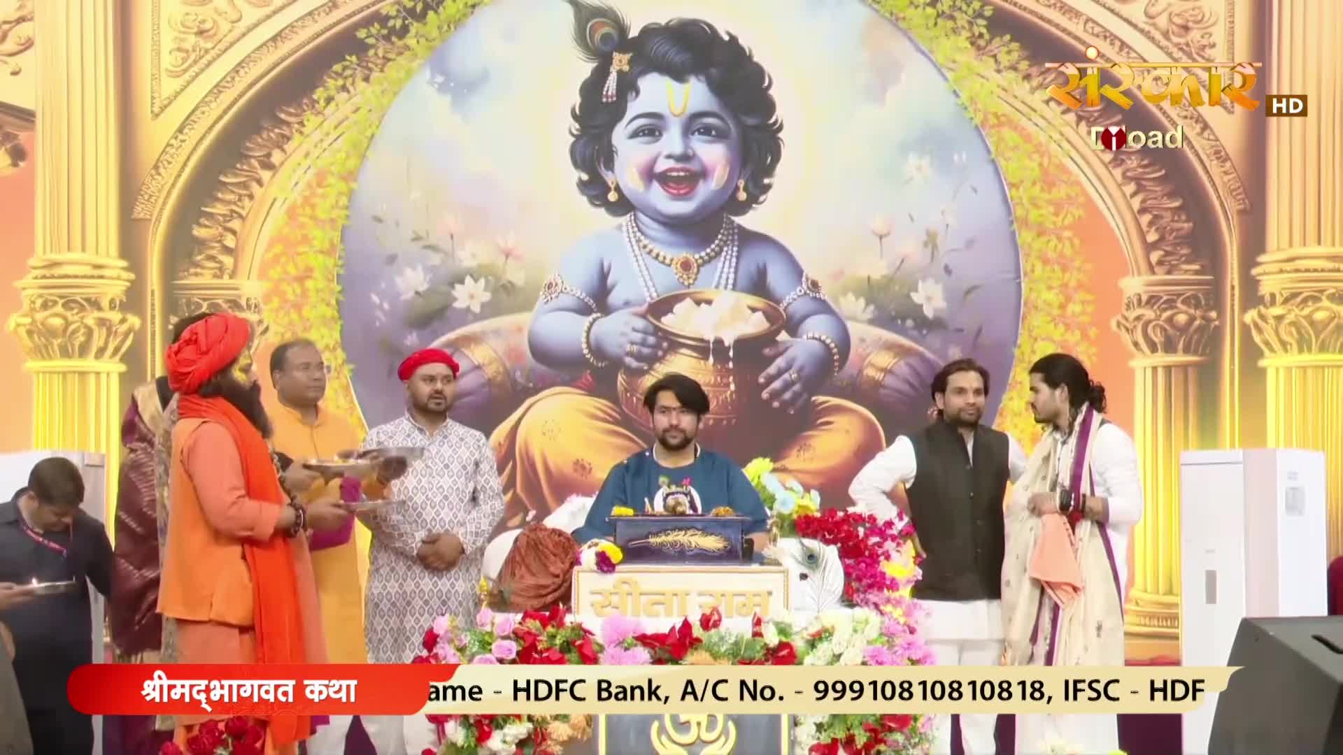 A man in an orange robe offers something on a plate to another man in a patterned shirt. Behind them, a large painting depicts a smiling baby Krishna holding a pot of butter.