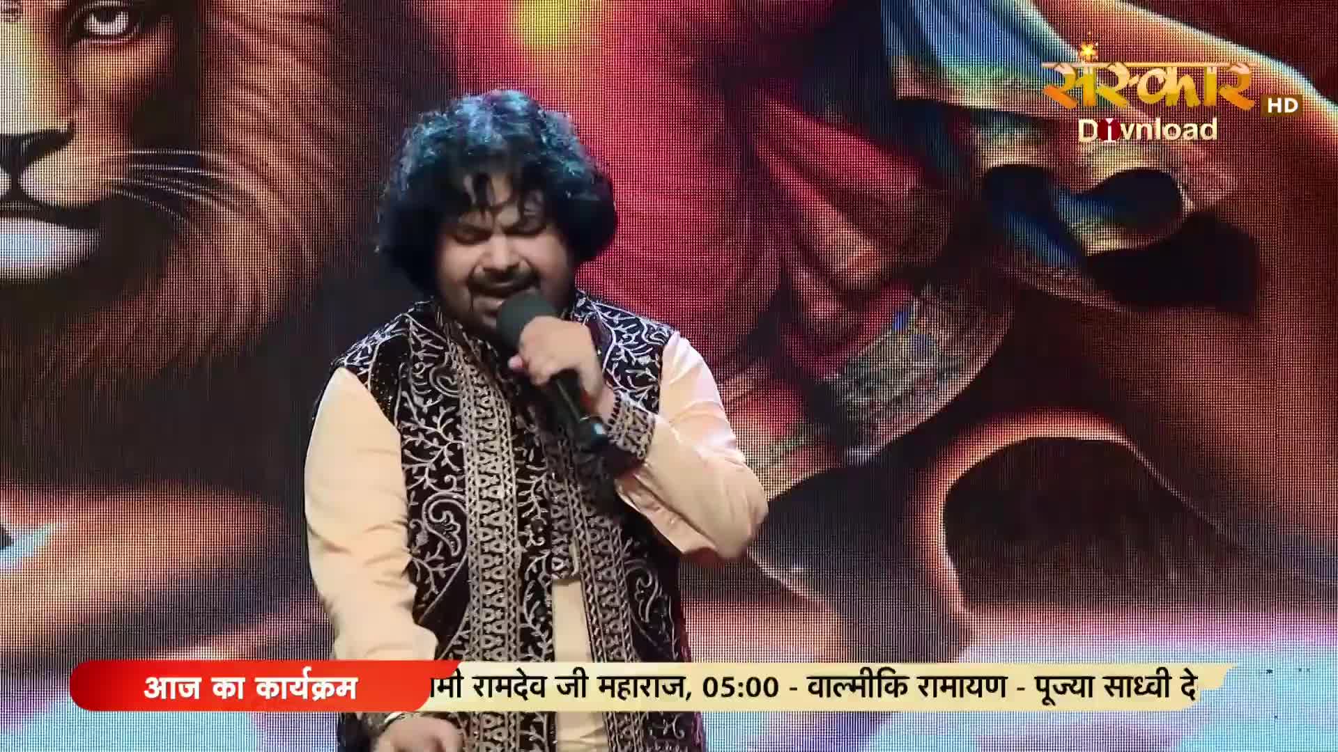 A man in a traditional Indian outfit sings into a microphone, his face lit by stage lights. Behind him, a large image of a lion's face dominates the backdrop.