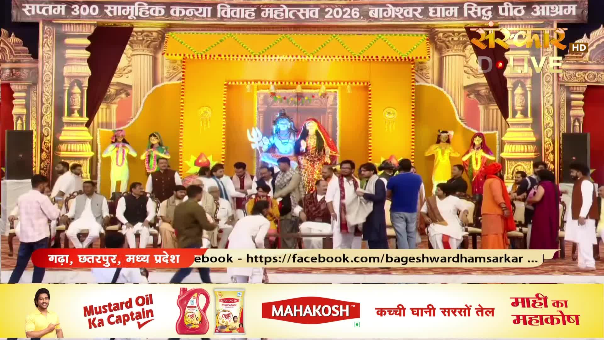 A large group of people are gathered on a stage for a wedding ceremony, with statues of deities visible behind them. A broadcast by Sanskar TV is in progress, documenting this event in India.