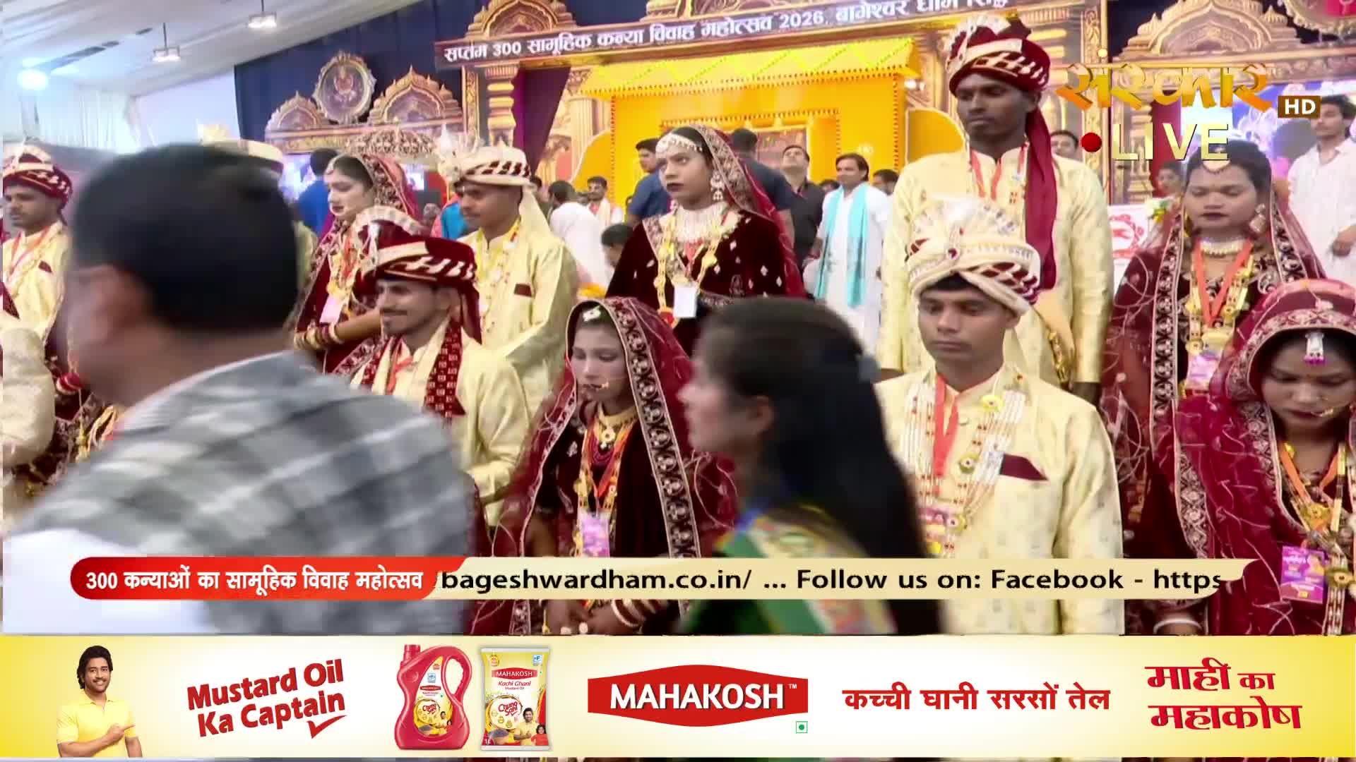 A procession of grooms in cream sherwanis and red turbans moves forward, each accompanied by a bride in a rich maroon sari and ornate jewelry. The air is filled with the sounds of a celebration, likely a mass wedding event broadcast on Sanskar TV.
