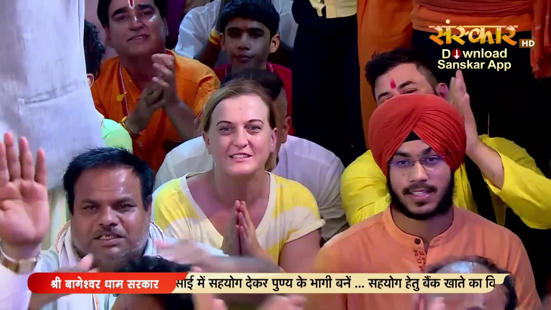 A woman with blonde hair smiles, her hands pressed together in prayer. Beside her, a young man in a bright orange turban looks forward, his gaze steady.