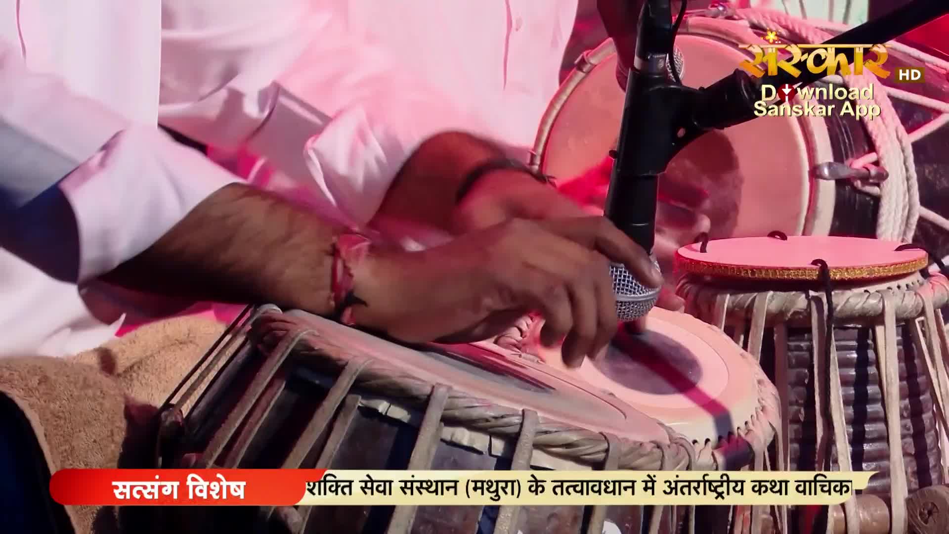 Hands move rapidly over the tabla drums, creating a rhythmic pulse. A microphone stands close, capturing the sound for Sanskar TV's broadcast.
