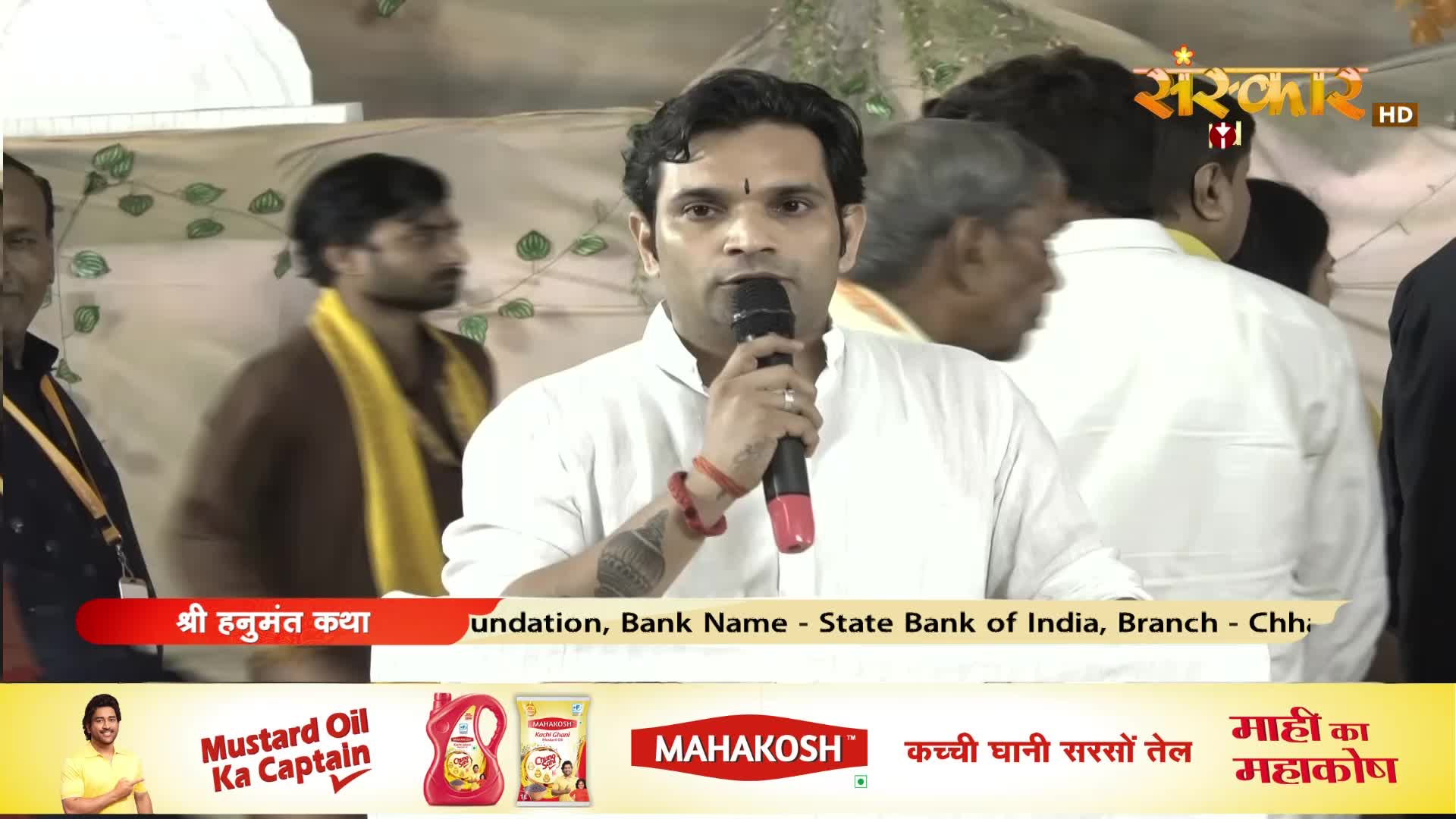 A man in a white kurta speaks into a microphone, his arm adorned with a red thread. Behind him, others in the crowd, some wearing yellow sashes, listen intently.