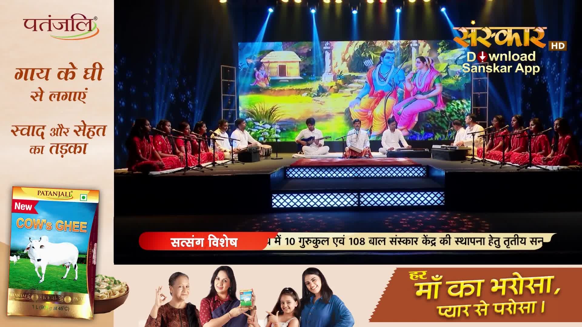 A group of women in red saris are singing on stage, accompanied by musicians playing traditional instruments. Behind them, a large screen displays a colorful depiction of Hindu deities.