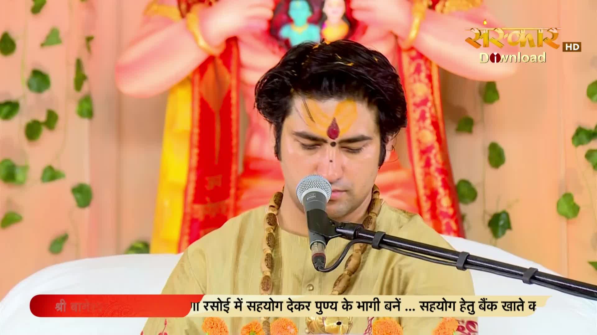 A man with a tilak on his forehead speaks into a microphone, his eyes downcast. Behind him, a large, colorful statue of a deity stands adorned with fabric.