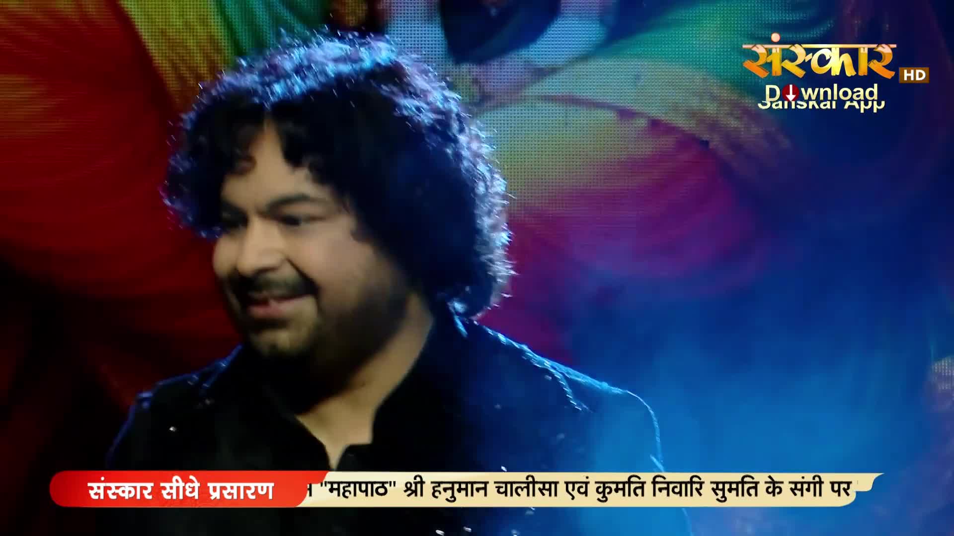 A man with curly dark hair smiles, his face illuminated by stage lights. The Sanskar TV logo appears in the upper right corner.
