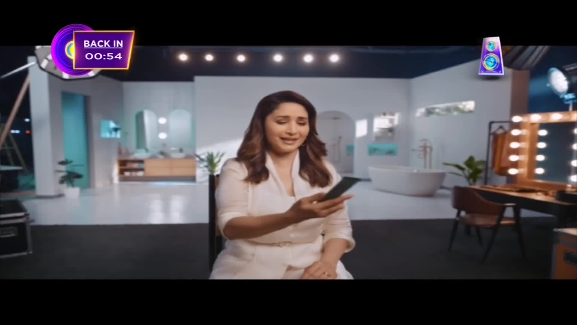 A woman in a white outfit sits holding a phone, looking at it with a slight smile. The set behind her is a modern, minimalist space with a bathtub and makeup vanity.
