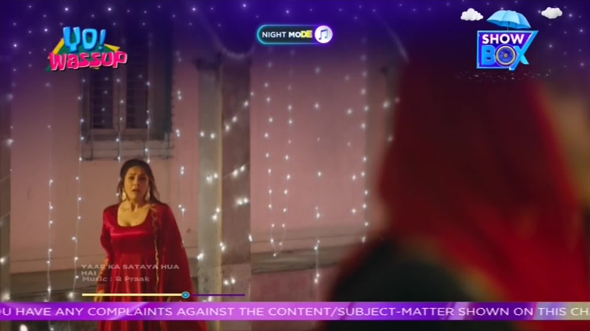 A woman in a red dress stands in front of a string of lights, singing on what appears to be a ShowBox program. The screen displays the "Yo! Wassup" logo, and the words "Yaay Ka Sataya Hua Hai" are visible.
