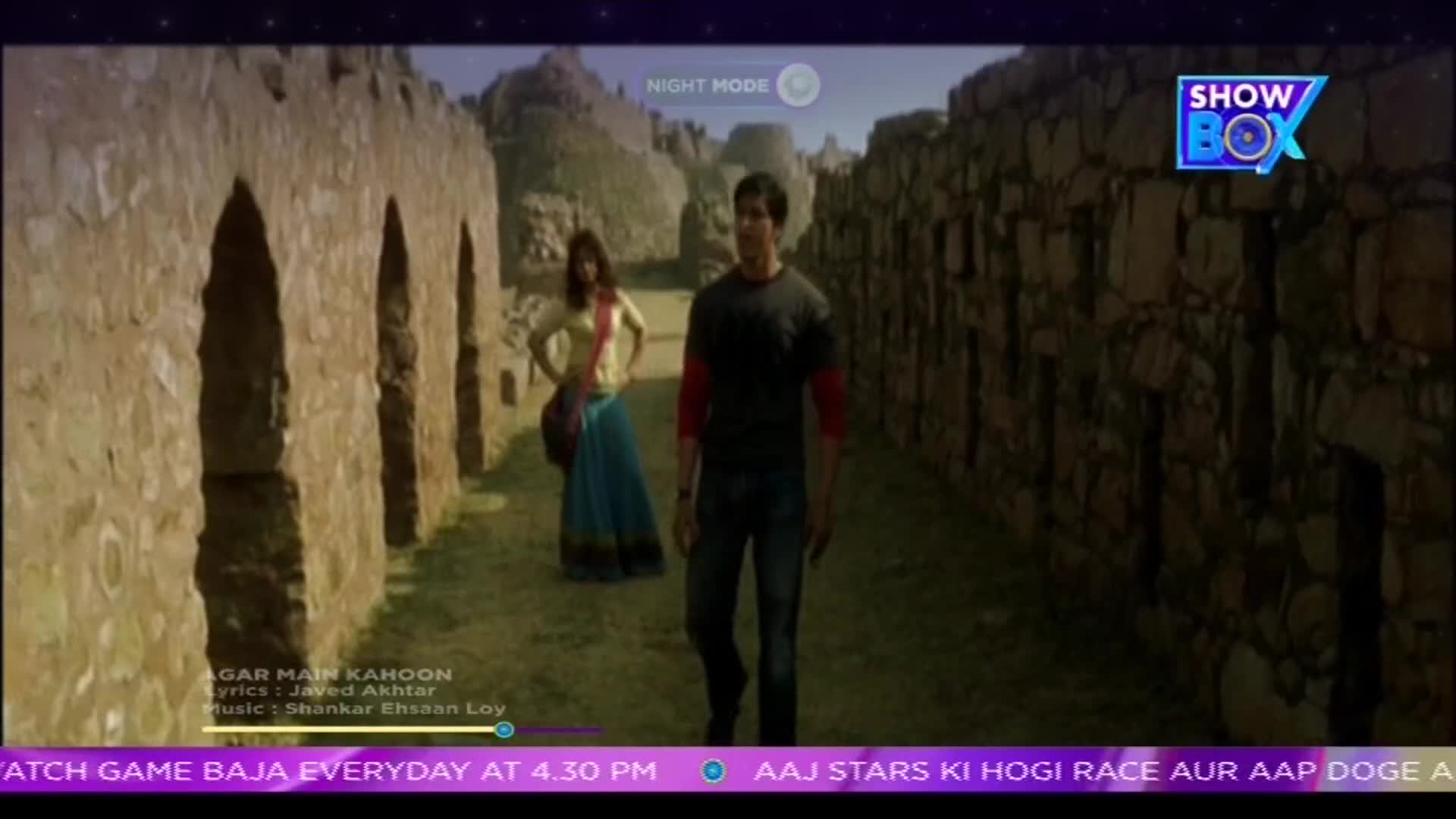 A man in a black tee walks towards the camera, while a woman in a colorful outfit stands behind him, posing. The scene, from what looks like an Indian music video on ShowBox, is framed by ancient stone walls.
