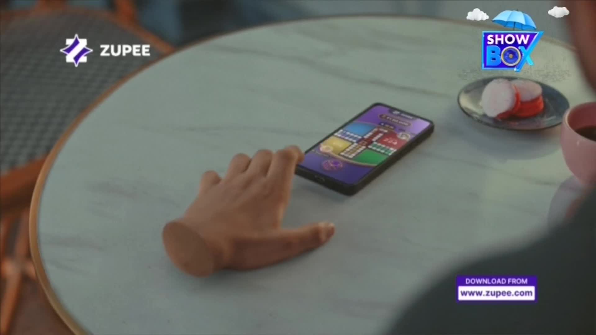 A hand hovers over a phone displaying a Ludo game, ready to make a move. Beside the phone, a small plate holds a couple of red and white sweets.
