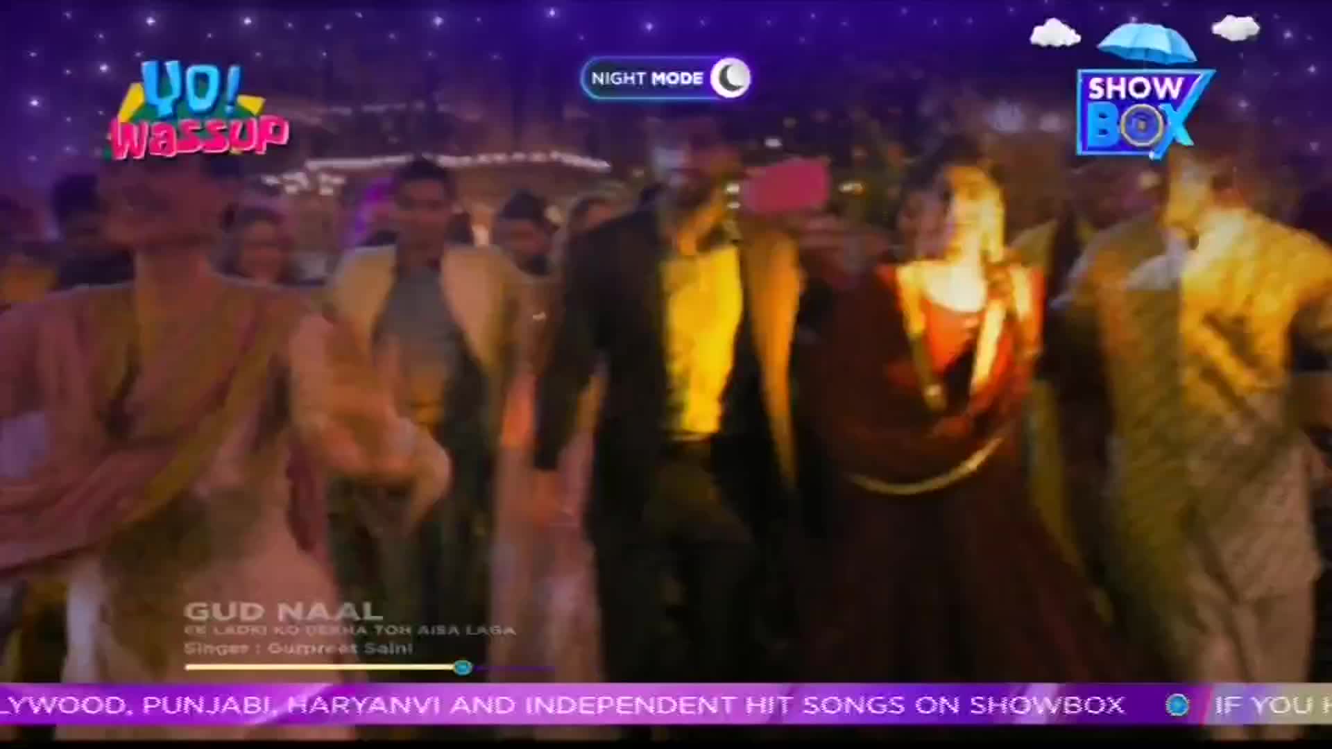People in brightly colored traditional clothing are dancing, moving to the music. The scene is from a music video on ShowBox, with a "Yo! Wassup" graphic in the corner.

