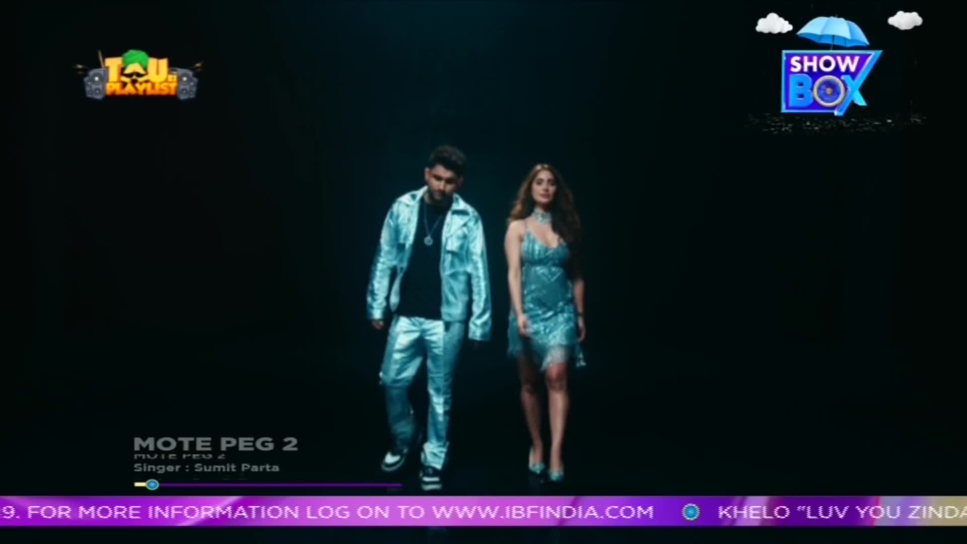 A man and woman, both in shimmering outfits, walk toward the camera against a dark background. The ShowBox logo and a music information banner are visible at the top and bottom of the screen.
