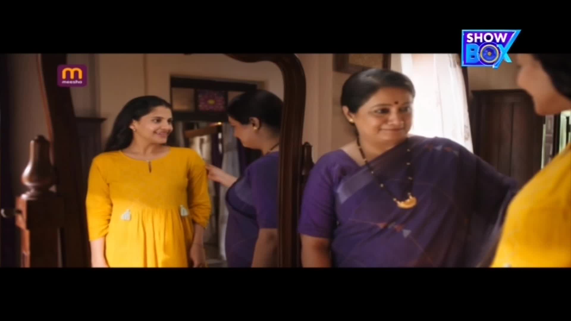 A woman in a yellow dress looks at herself in the mirror while another woman in a purple sari adjusts her outfit. The woman in the purple sari smiles, seemingly pleased with the result.

