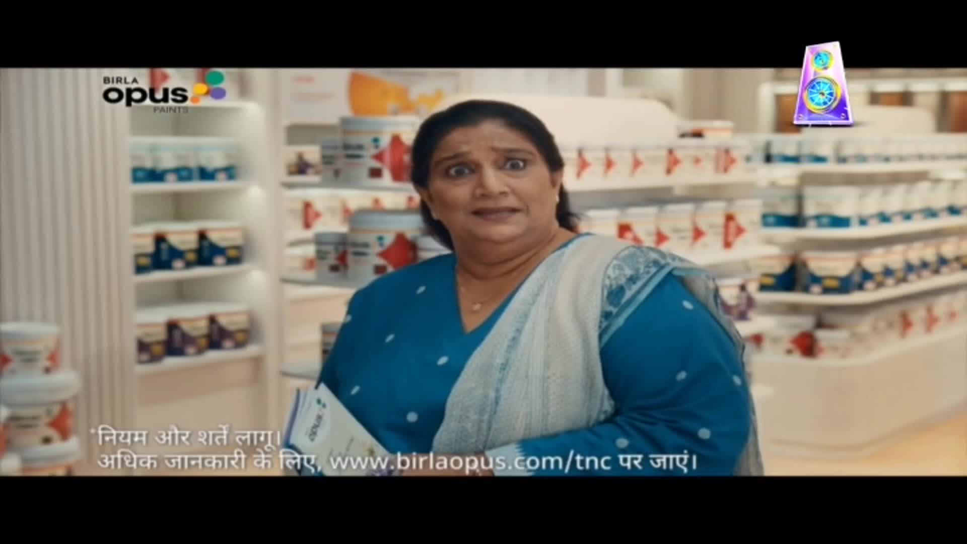 A woman in a blue sari is holding a pamphlet and looking surprised in a paint store. Shelves of paint cans line the background, and a colorful logo for Birla Opus Paints is visible.
