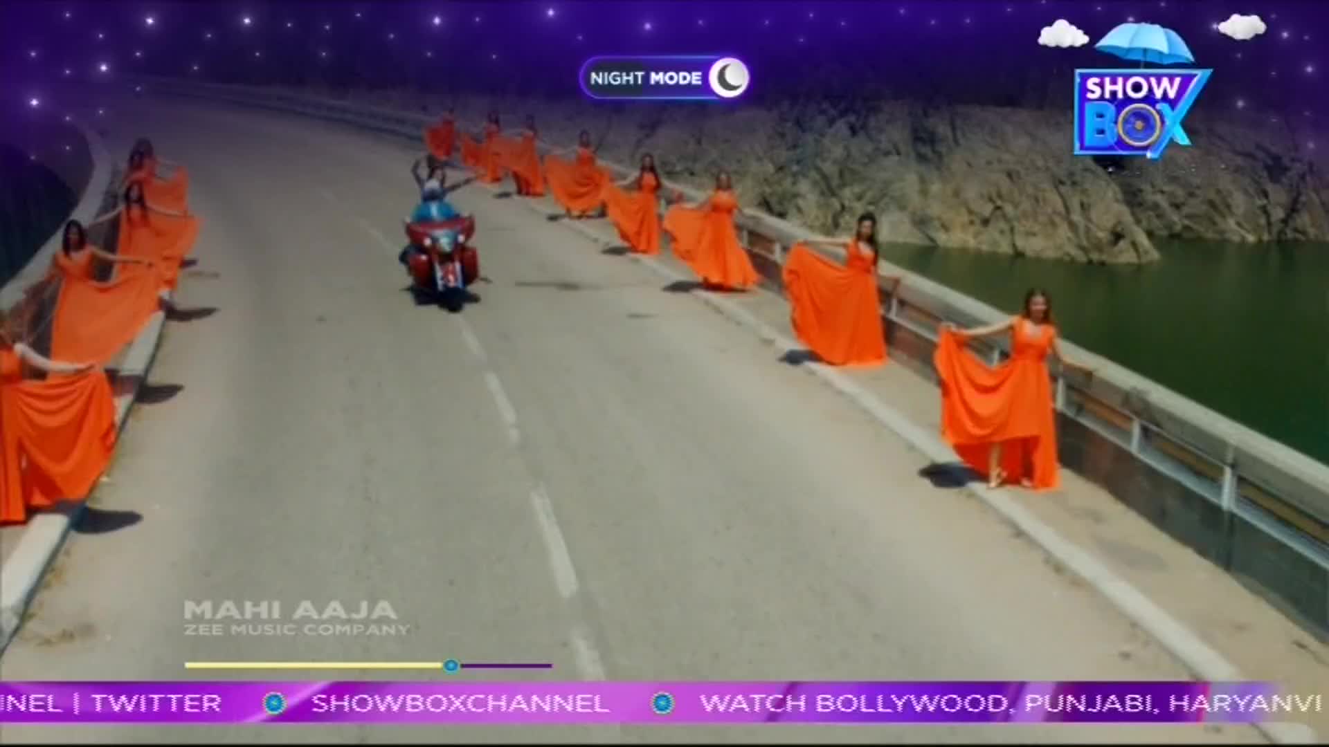 A motorcycle speeds down the road, flanked by women in bright orange dresses. They hold the fabric out as if dancing, while the ShowBox logo sits in the corner.
