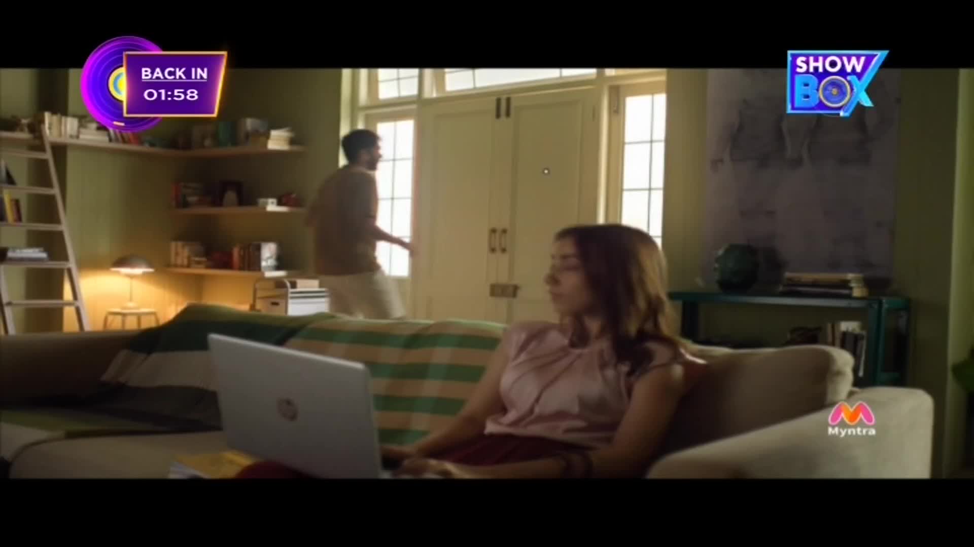 A man walks away from the camera towards a brightly lit doorway. A woman sits on a sofa, looking down at her laptop. A man walks away from the camera towards a brightly lit doorway. A woman sits on a sofa, looking down at her laptop.