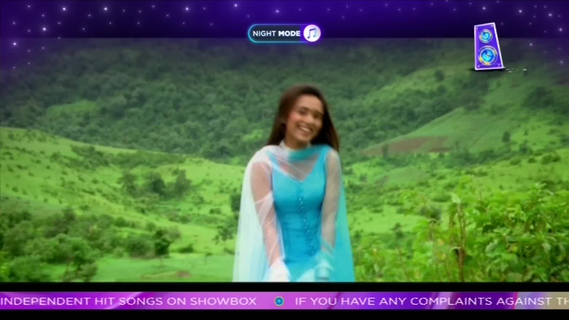 A young woman in a bright blue salwar kameez smiles and laughs, her sheer dupatta flowing around her. The lush green hills of India form a vibrant backdrop to this scene, a moment captured from a ShowBox music video. A young woman in a bright blue salwar kameez smiles and laughs, her sheer dupatta flowing around her. The lush green hills of India form a vibrant backdrop to this scene, a moment captured from a ShowBox music video.