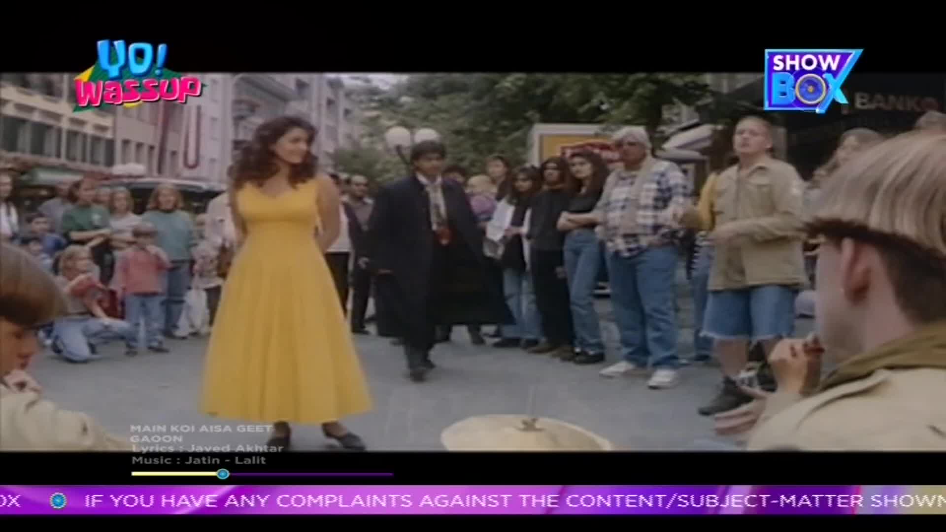A woman in a bright yellow dress twirls as a man in a long black coat walks past her. A crowd of onlookers watches the scene unfold on a European street, likely as part of a Bollywood production for ShowBox. A woman in a bright yellow dress twirls as a man in a long black coat walks past her. A crowd of onlookers watches the scene unfold on a European street, likely as part of a Bollywood production for ShowBox.