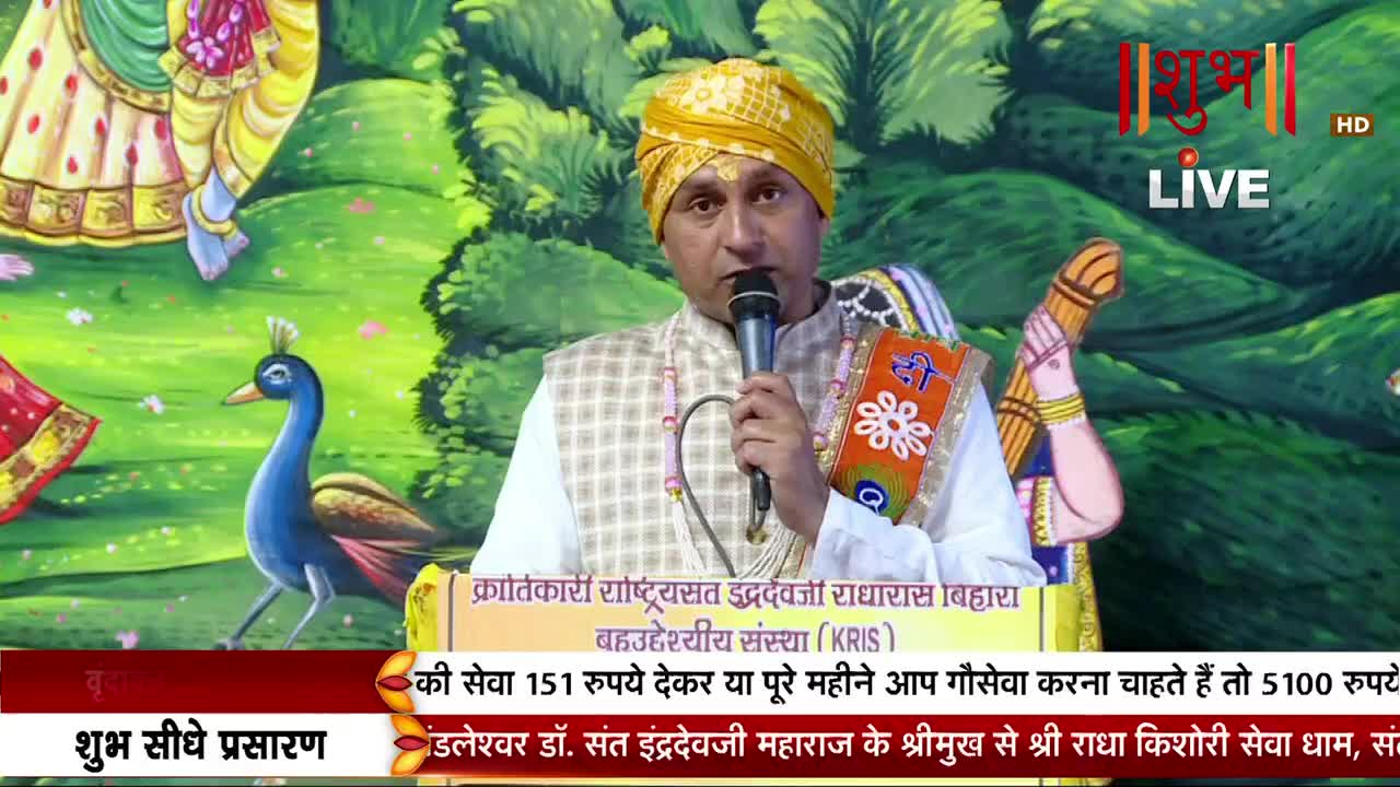 A man in a yellow turban speaks into a microphone, with a painted peacock and figures in the background. A banner at the bottom of the screen displays text in Hindi.