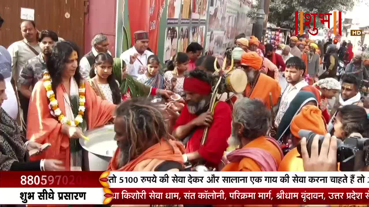 A man in an orange robe, adorned with a floral garland, offers a bowl of white liquid to a man with long, matted hair. A crowd of people, many in saffron and orange attire, gathers around them, some holding musical instruments.
