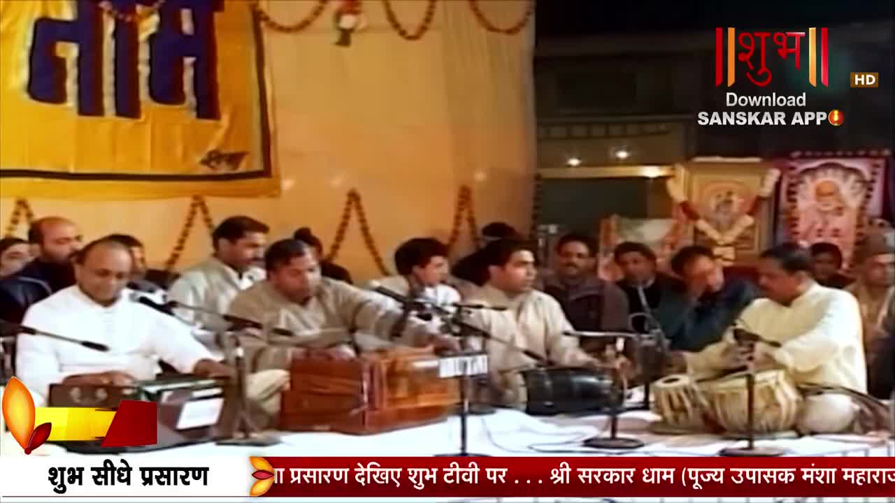 A man in a white kurta plays a harmonium, his fingers moving across the keys. Beside him, another man taps out a rhythm on a tabla, his hands a blur against the drumheads.