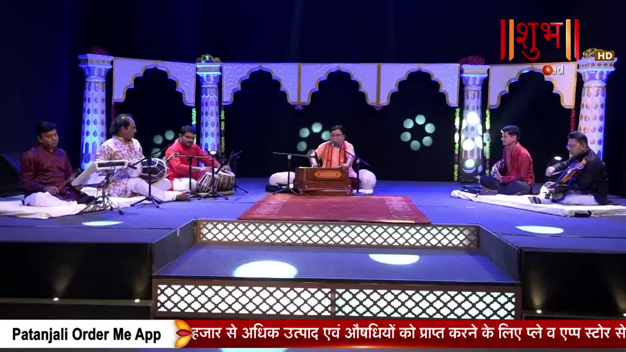 A group of musicians sits on a stage, performing Indian classical music. One man plays a harmonium while others accompany him on tabla and string instruments. The Shubh TV logo is visible in the background.