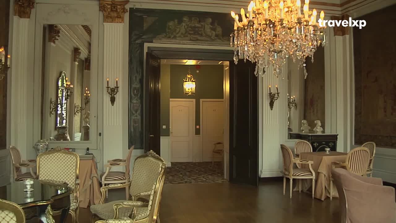 A grand chandelier hangs above tables set for a meal, reflecting in a large mirror. Through a doorway, two white doors lead to another space, lit by a hanging lamp.
A grand chandelier hangs above tables set for a meal, reflecting in a large mirror. Through a doorway, two white doors lead to another space, lit by a hanging lamp.