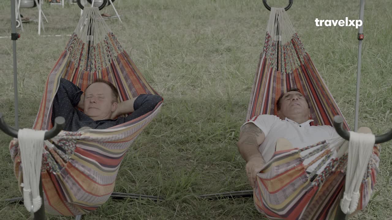 Two men are relaxing in hammocks, their eyes closed, with arms behind their heads. The colorful striped fabric of the hammocks contrasts with the green grass around them.
Two men are relaxing in hammocks, their eyes closed, with arms behind their heads. The colorful striped fabric of the hammocks contrasts with the green grass around them.