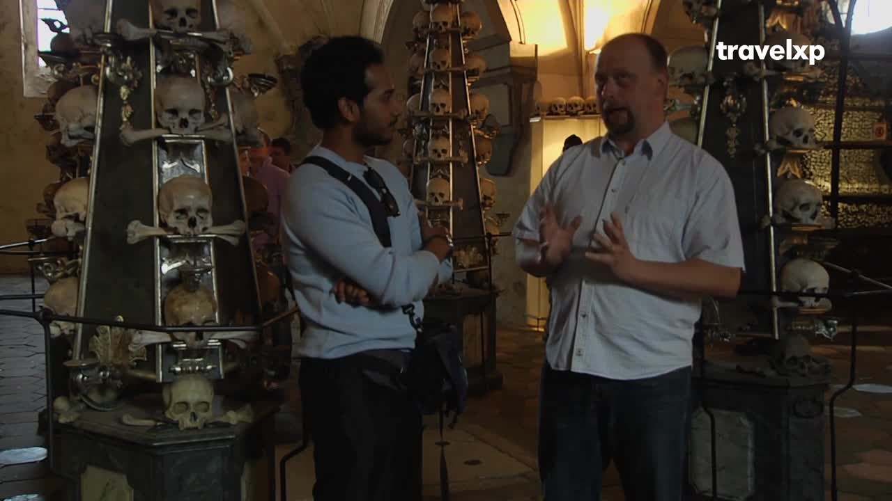 A man in a light blue shirt gestures with both hands, explaining something to the other man, who stands with his arms crossed. Behind them, skulls and bones are arranged on tall, ornate structures.
A man in a light blue shirt gestures with both hands, explaining something to the other man, who stands with his arms crossed. Behind them, skulls and bones are arranged on tall, ornate structures.