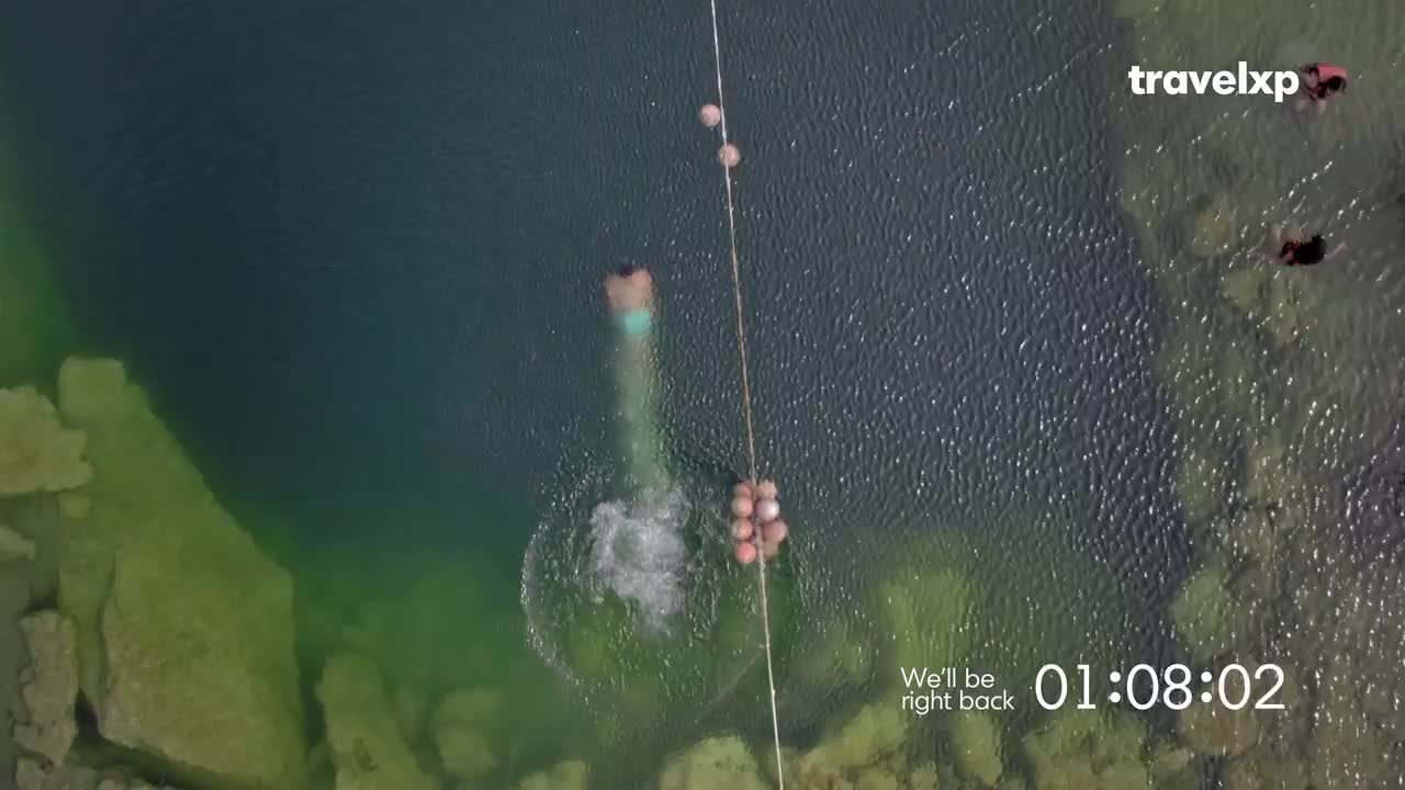 A person plunges into the clear water, creating a splash. Nearby, others swim in the shallow, rocky area. A person plunges into the clear water, creating a splash. Nearby, others swim in the shallow, rocky area.