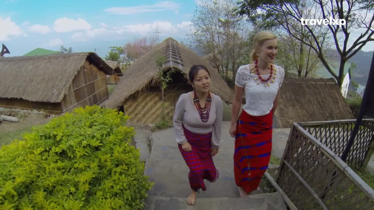 Two women, one in traditional Indian attire, descend stone steps past thatched-roof huts. Their colorful woven skirts stand out against the lush green foliage. Two women, one in traditional Indian attire, descend stone steps past thatched-roof huts. Their colorful woven skirts stand out against the lush green foliage.
