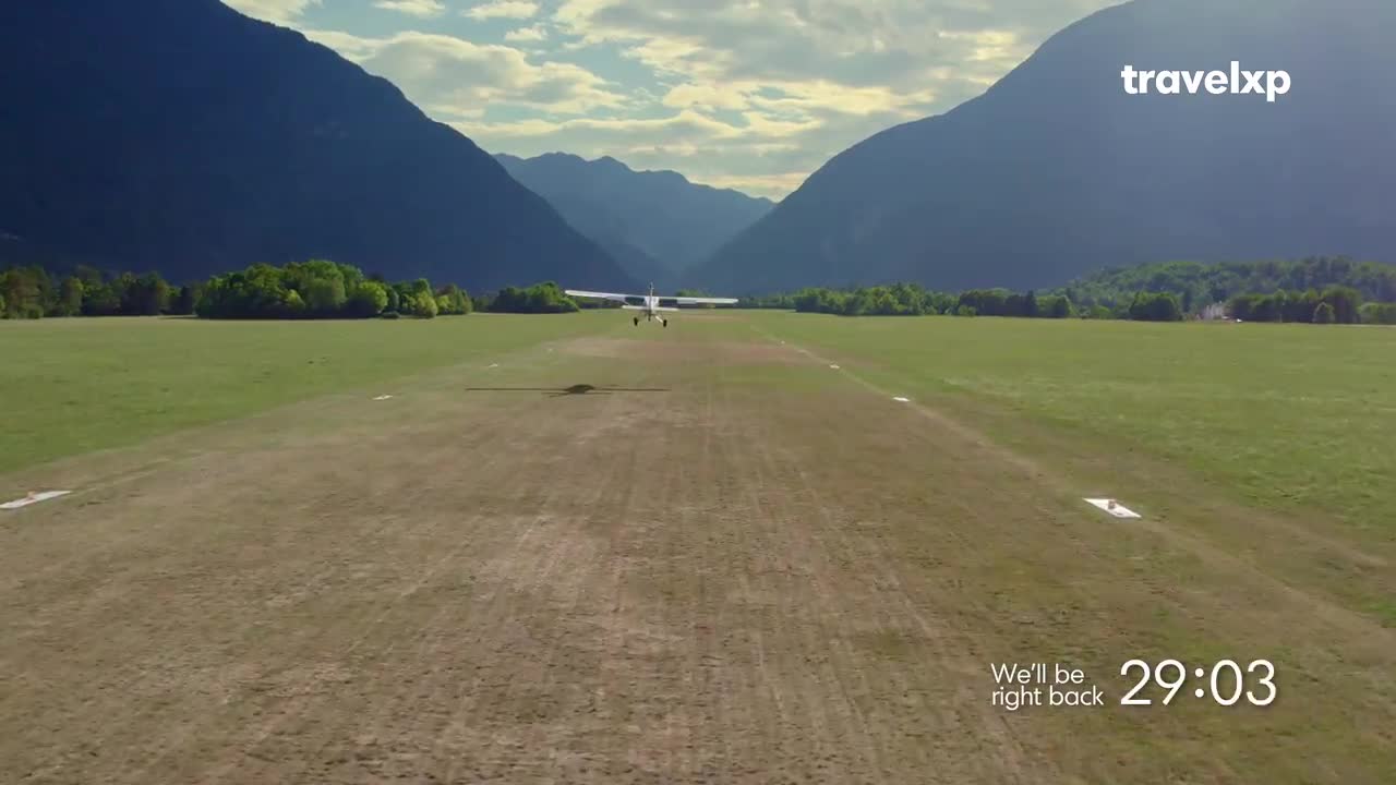 A small plane is lifting off from a grassy runway. The Travelxp logo appears in the corner, hinting at an upcoming travel segment. A small plane is lifting off from a grassy runway. The Travelxp logo appears in the corner, hinting at an upcoming travel segment.