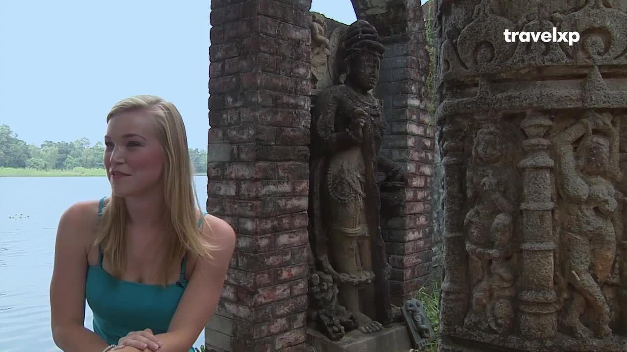 A woman with blonde hair sits by a tranquil lake, her gaze directed off-camera. Behind her, ancient stone carvings and weathered brickwork stand as silent witnesses to India's rich history. A woman with blonde hair sits by a tranquil lake, her gaze directed off-camera. Behind her, ancient stone carvings and weathered brickwork stand as silent witnesses to India's rich history.