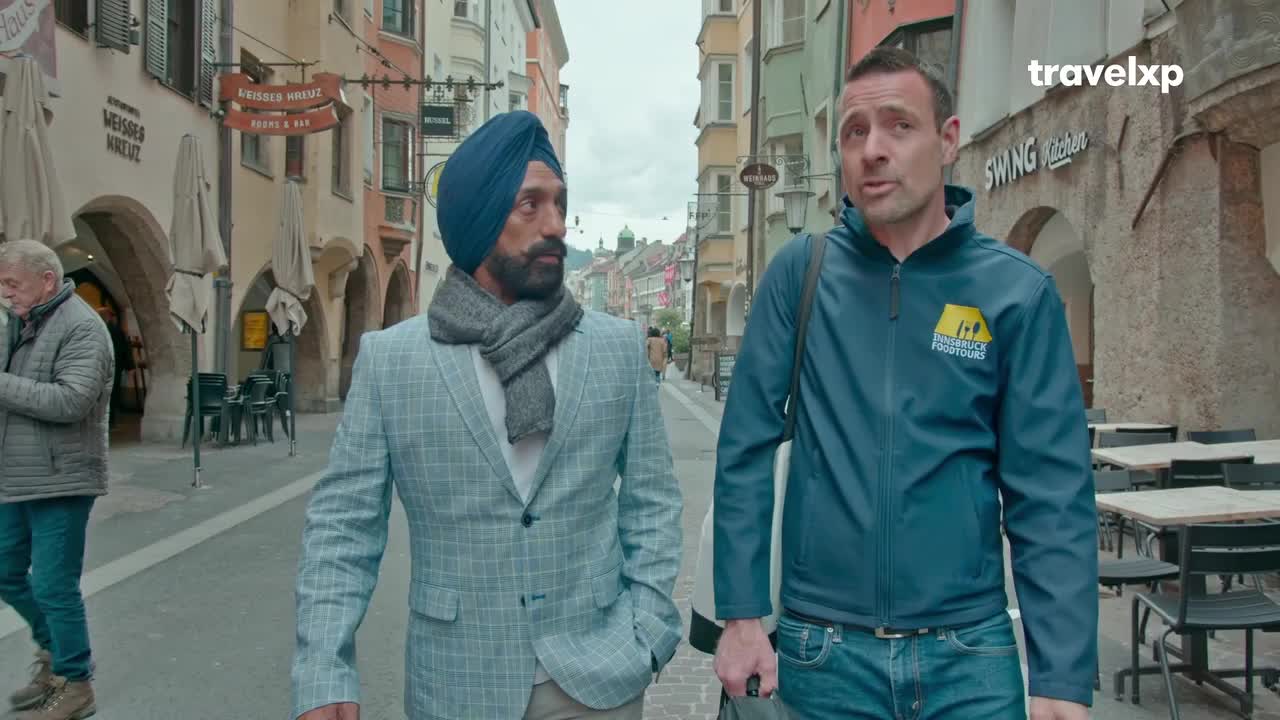 Two men walk down a cobblestone street lined with shops. The man on the left wears a light blue checkered suit and a grey scarf, while the man on the right sports a dark blue jacket with a Travelxp logo. Two men walk down a cobblestone street lined with shops. The man on the left wears a light blue checkered suit and a grey scarf, while the man on the right sports a dark blue jacket with a Travelxp logo.