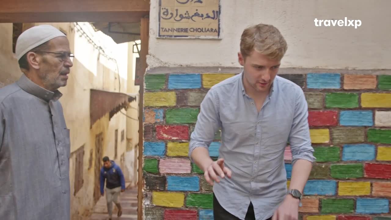 A man in a light blue shirt gestures animatedly beside a vibrantly painted wall. Further down the narrow alley, another man in a blue jacket walks away. A man in a light blue shirt gestures animatedly beside a vibrantly painted wall. Further down the narrow alley, another man in a blue jacket walks away.