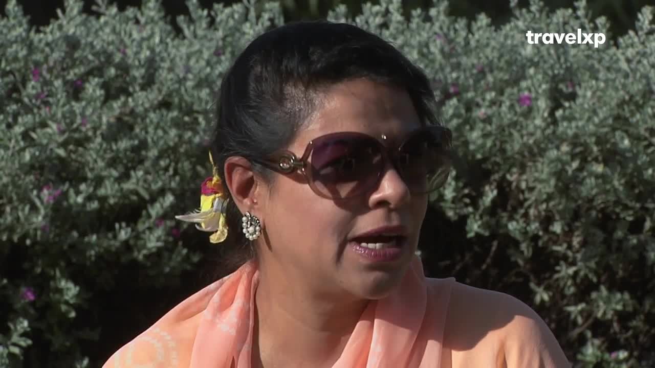 A woman with a bright yellow hair accessory speaks, her sunglasses reflecting the sun. The Travelxp logo is visible in the upper right corner. A woman with a bright yellow hair accessory speaks, her sunglasses reflecting the sun. The Travelxp logo is visible in the upper right corner.