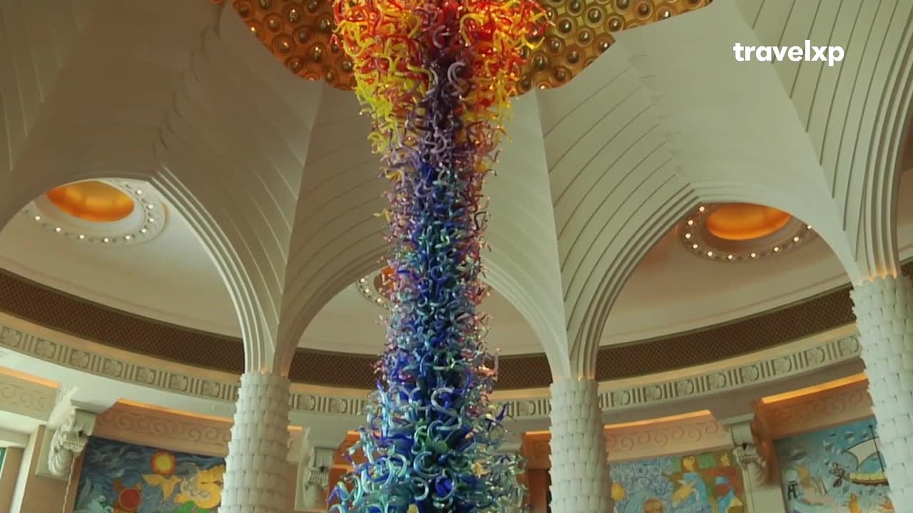 A towering glass sculpture, shimmering with colors from yellow to deep blue, hangs from the ceiling. The intricate artwork is set within a grand, arched interior, hinting at the opulent settings often featured on Travelxp. A towering glass sculpture, shimmering with colors from yellow to deep blue, hangs from the ceiling. The intricate artwork is set within a grand, arched interior, hinting at the opulent settings often featured on Travelxp.