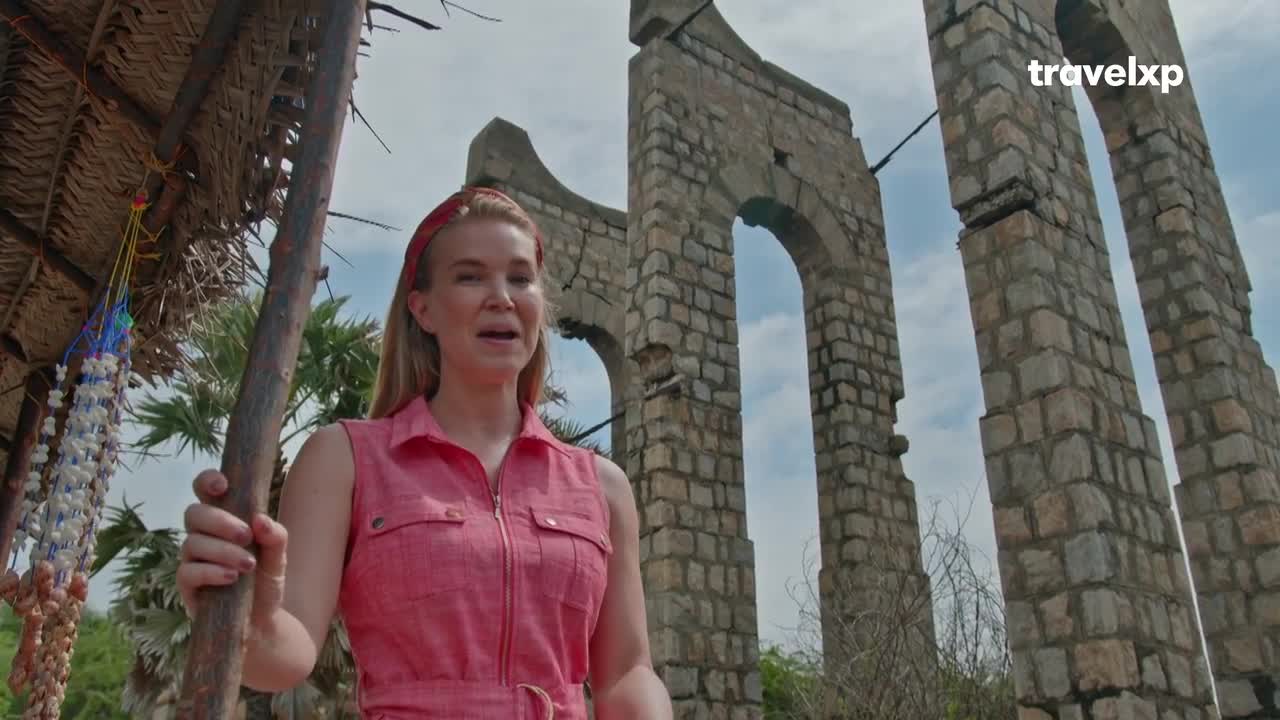 A woman in a pink dress stands near a thatched shelter, speaking. Behind her, the crumbling stone arches of an ancient structure reach towards the sky. A woman in a pink dress stands near a thatched shelter, speaking. Behind her, the crumbling stone arches of an ancient structure reach towards the sky.