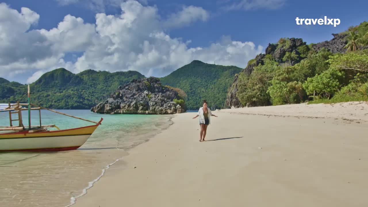 A woman walks along a white sand beach, her arms outstretched. A traditional boat rests in the shallows of the turquoise water, its yellow hull gleaming. A woman walks along a white sand beach, her arms outstretched. A traditional boat rests in the shallows of the turquoise water, its yellow hull gleaming.