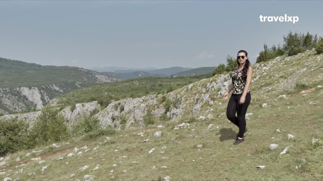 A woman walks across a grassy hillside, her black leggings and patterned top a stark contrast to the rugged landscape. Behind her, a vast valley unfolds, its rocky slopes covered in green foliage under a clear blue sky. A woman walks across a grassy hillside, her black leggings and patterned top a stark contrast to the rugged landscape. Behind her, a vast valley unfolds, its rocky slopes covered in green foliage under a clear blue sky.