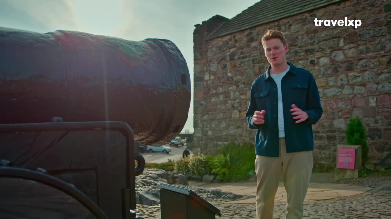 A young man stands beside a large, dark cannon, gesturing as he speaks. Behind him, a stone building rises against a bright sky, with a few cars visible in the distance. A young man stands beside a large, dark cannon, gesturing as he speaks. Behind him, a stone building rises against a bright sky, with a few cars visible in the distance.