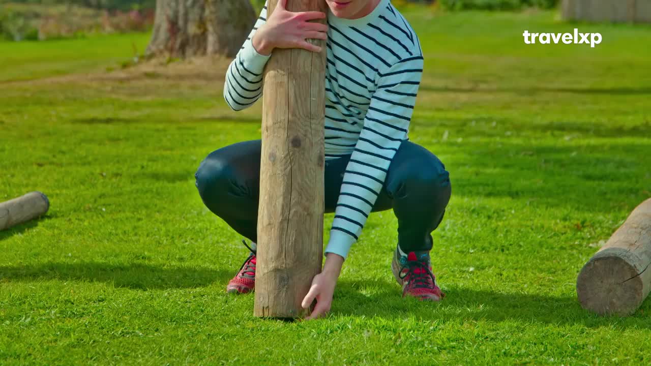 A man in a striped sweater crouches, his hands gripping a thick wooden post. Another log lies on the grass nearby, suggesting a game or activity. A man in a striped sweater crouches, his hands gripping a thick wooden post. Another log lies on the grass nearby, suggesting a game or activity.