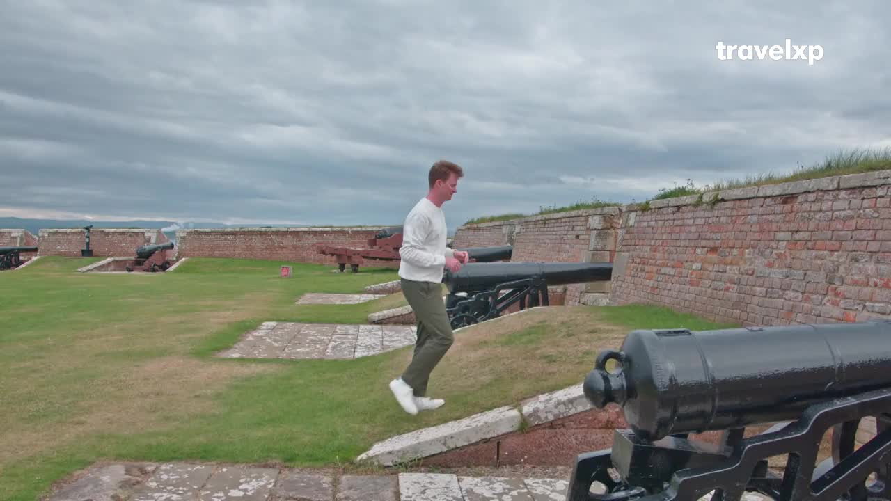 A man in a white sweater and green pants bounds across the grassy ramparts of an old fort. Rows of dark cannons sit ready along the brick walls, a testament to history. A man in a white sweater and green pants bounds across the grassy ramparts of an old fort. Rows of dark cannons sit ready along the brick walls, a testament to history.