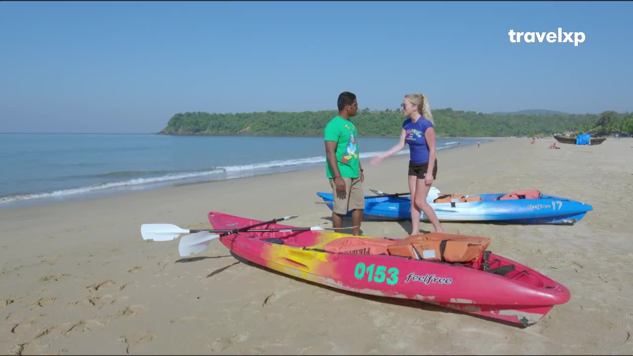 A man in a green t-shirt and a woman in a blue top stand by kayaks on a sandy Indian beach. The kayaks, one bright pink and the other blue, are ready for use near the gentle waves.