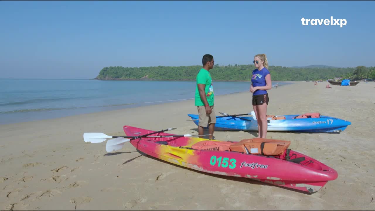 Two people stand on a sandy beach in India, discussing kayaks. The bright pink and yellow kayak in the foreground has paddles resting across it.