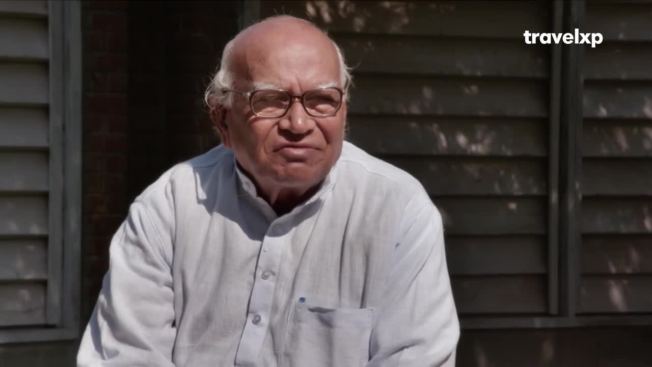An elderly gentleman with glasses sits outdoors, his white shirt catching the sunlight. The Travelxp logo appears in the upper right corner.
