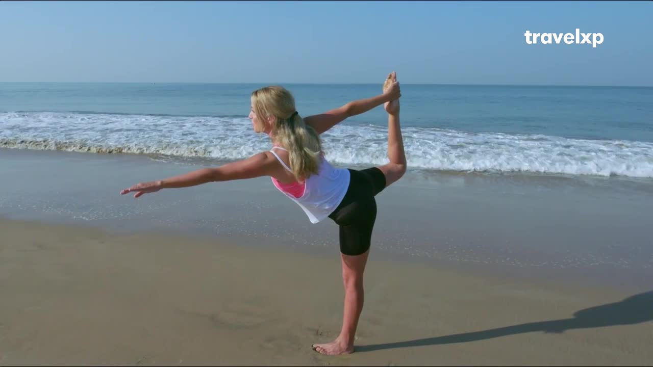 A woman balances on one leg, her other foot raised and held by her hand. Gentle waves lap the shore behind her, a scene perfect for a Travelxp segment on India's coastline.
